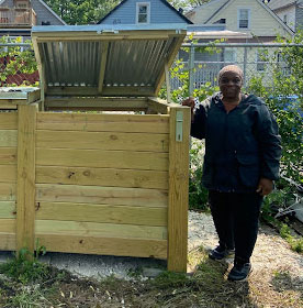 A person stands outdoors next to a large wooden compost bin with the lid lifted