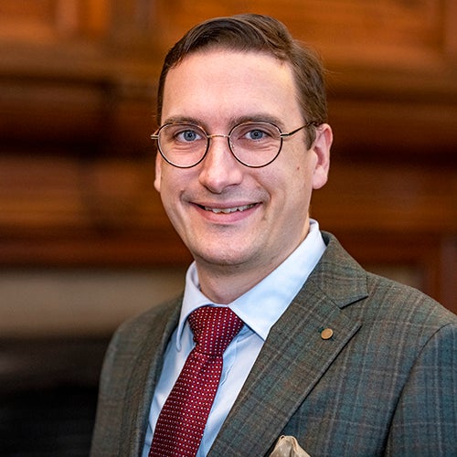 Person smiling with short brown hair and glasses, wearing a suit and tie