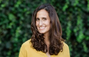 Dr. Priya Fielding-Singh is pictured from the shoulders up wearing a yellow sweater standing in front of a green background. She looks into the camera smiling.