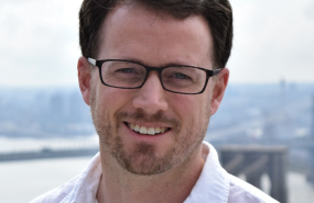 Scott MacMillan is pictured from the shoulders up, wearing a white button-down shirt and black glasses. He is looking into the camera and smiling.