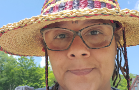 Ife Kilimanjaro is pictured from the shoulders up, wearing a light gray shirt, striped hat, and glasses as she smiles into the camera.