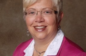 Jacqueline Applegate is pictured from the shoulders up wearing a pink sweater over a white button-up shirt, smiling into the camera.