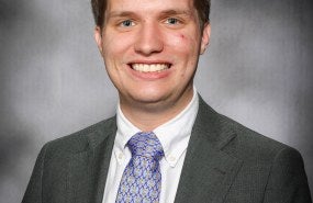Clayton is pictured from the shoulders up, smiling into the camera wearing a suit and tie.