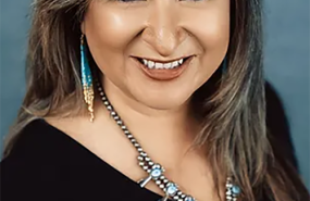 Leah Altman is pictured from the shoulders up smiling into the camera and wearing a black shirt with a necklace and earrings.