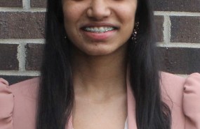 Avani Rai is pictured from the shoulders up wearing a pink blazer over a white shirt, smiling into the camera.