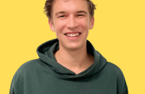 Ben Collier is pictured from the shoulders up wearing a green sweatshirt, smiling into the camera and standing in front of a yellow background.