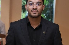 Abdullah Al Maruf is pictured from the waist up, looking into the camera, wearing a black suit jacket over a black button-up shirt.
