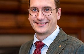 Person smiling with short brown hair and glasses, wearing a suit and tie