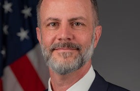 Portrait of Kim Ruhl wearing a suit with a grey tie, in front of the American flag
