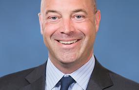 Headshot of Chris Keogh smiling and wearing dark grey jacket, dark blue tie in front of blue background