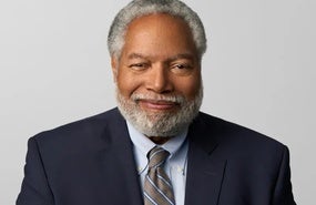 Headshot of Lonnie Bunch smiling, wearing black jacket and grey tie in front of grey background
