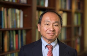 Headshot of Francis Fukuyama wearing black blazer, red tie, blue shirt in front of bookshelves