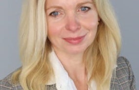 Headshot of Margaret Kruk wearing grey plaid blazer, smiling, in front of grey background