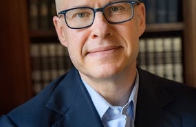 Headshot of Max Boot wearing navy suit jacket and glasses in front of bookshelf