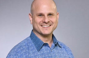 Headshot of Dean Karlan smiling in front of grey background with blue shirt
