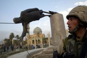 A statue of Saddam Hussein falling off its pedestal with a person in a military uniform watching. 