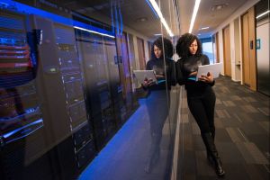 A woman in all black stands next to servers, behind glass, holding a laptop computer