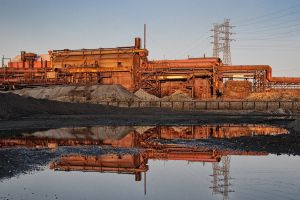 An exterior shot of a factory in the rust belt at sunset, with a puddle of water in the foreground