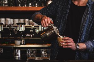A barista pours coffee from a silver pitcher into a cup