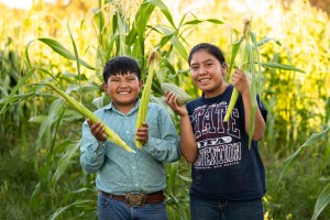 Doyan and Kaiesta smile into the camera as they hold up two stalks of corn.