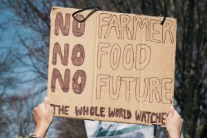 A woman holds up a sign at a protest that says, "no farmer, no food, no future; the whole world watches."