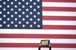 a person holds a sign that says vote in front of an American flag