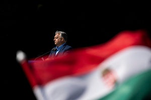 Hungarian Prime Minister Viktor Orbán stands at a podium with a microphone as a Hungarian flag waves in the foreground.