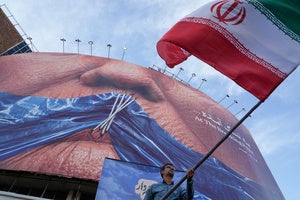 A man waves an Iranian flag under a billboard with graphic showing Strait of Hormuz and sewn lips of USPresident Donald Trump