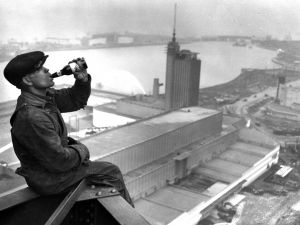 An iron worker sits on the edge of a skyscraper while drinking a beverage