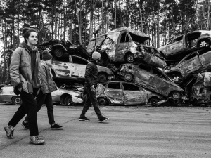 People walking through destruction in Ukraine, with stacks of destroyed cars in the background
