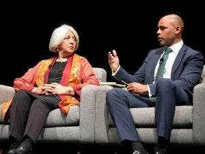 Two people sit in chairs, talking on stage