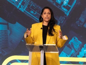 A person wearing a yellow shirt speaks into a microphone on-stag behind a podium