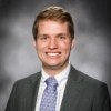Clayton is pictured from the shoulders up, smiling into the camera wearing a suit and tie.