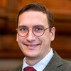 Person smiling with short brown hair and glasses, wearing a suit and tie