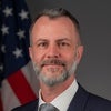 Portrait of Kim Ruhl wearing a suit with a grey tie, in front of the American flag