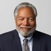 Headshot of Lonnie Bunch smiling, wearing black jacket and grey tie in front of grey background