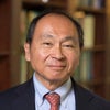 Headshot of Francis Fukuyama wearing black blazer, red tie, blue shirt in front of bookshelves
