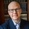 Headshot of Max Boot wearing navy suit jacket and glasses in front of bookshelf