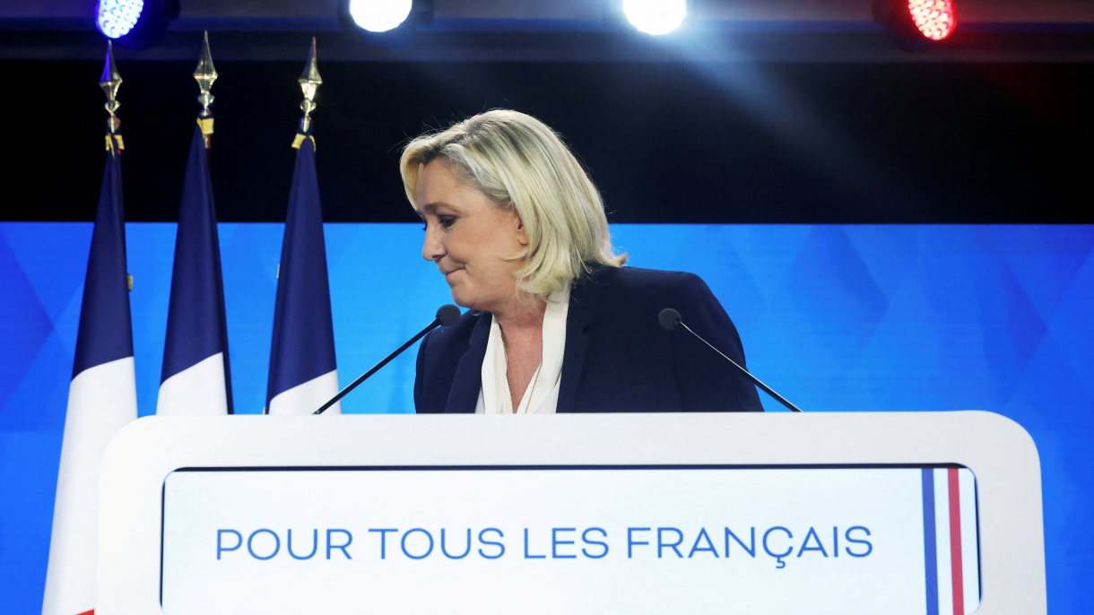 Marine Le Pen in front of a podium looking to her right with French flags.