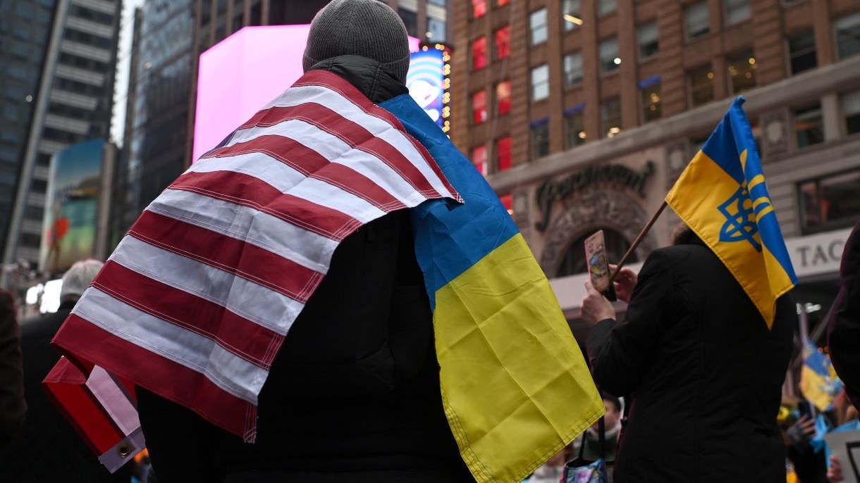 Person seen from behind with an American flag and Ukraine flag draped over their shoulders, grey beanie on their head.