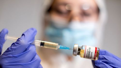A woman holds a small bottle labeled with a "Coronavirus COVID-19 Vaccine" sticker and a medical syringe in this illustration