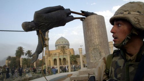 A statue of Saddam Hussein falling off its pedestal with a person in a military uniform watching. 