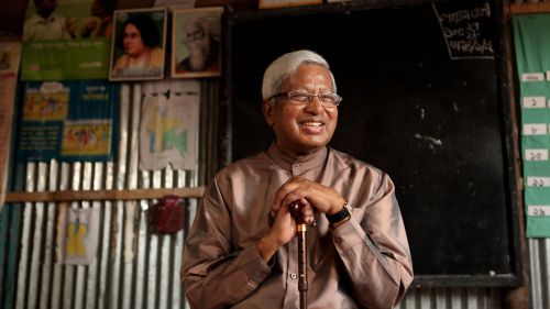 Sir Fazle Hasan Abed is sitting in a classroom in front of a black board, smiling and looking to the right as he holds a brown cane.