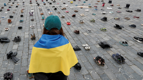 A woman draped in the Ukrainian flag looks at shoes symbolizing war crimes committed against Ukrainian civilians