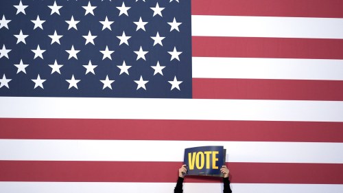 a person holds a sign that says vote in front of an American flag