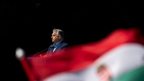Hungarian Prime Minister Viktor Orbán stands at a podium with a microphone as a Hungarian flag waves in the foreground.