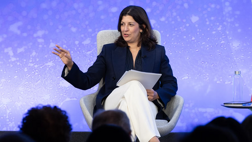 Leslie Vinjamuri talking to an audience in white pants, navy blazer