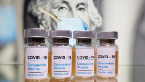 Vials with a sticker reading, "COVID-19 / Coronavirus vaccine / Injection only" are seen in front of a displayed U.S. dollar banknote with George Washington and a printed mask