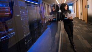 A woman in all black stands next to servers, behind glass, holding a laptop computer