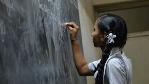 A young Indian girl writes on a chalkboard in a classroom